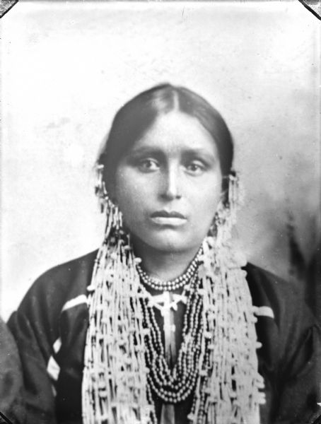 A studio portrait of a Ho-Chunk woman. She wears drop earrings and many beaded necklaces.
