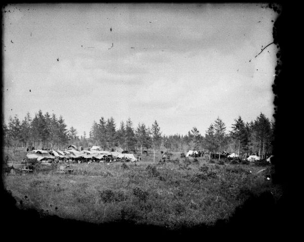 View from field of a Ho-Chunk encampment in a field surrounded by trees, probably a powwow and/or medicine dance site.
