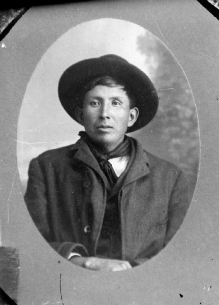 Copy photograph of an oval-framed waist-up studio portrait of a Ho-Chunk man posing sitting in front of a painted backdrop wearing a winter coat, bandana, and hat. Identified as Jerome Baptiste (Wakonhonka, King of Snakes) or St. Cyr of Nebraska.