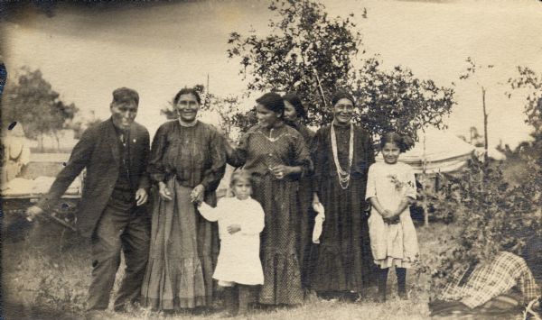 A group of four Ho-Chunk women, one Ho-Chunk man, and two Ho-Chunk girls posed standing outdoors in front of some shrubs and a tent.
