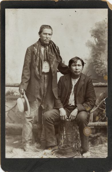 Studio portrait of two Ho-Chunk men in front of a painted backdrop and a prop wooden fence. The man standing on the left is wearing an untied bandana around his neck and is holding a hat in his right hand. The man on the right is seated on a stump and his hat is on a rug in front of him.