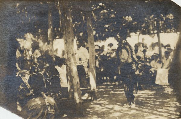 A Ho-Chunk gathering, possibly a powwow. Several Ho-Chunk men are wearing regalia, including feathered headdresses and bandoleers. They are under a structure supported by wood posts, and a group of people are sitting in the background watching the men.