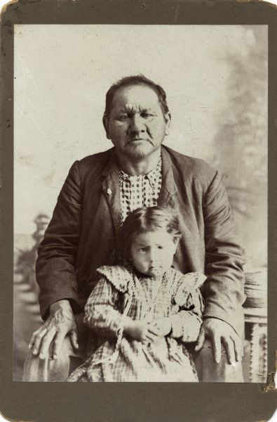 A studio portrait of a Ho-Chunk man and girl in front of a painted backdrop. The man is wearing a suit coat with a checked shirt underneath and is posed sitting behind the girl, who is wearing a dress and a necklace. Frank Mike, seated with daughter who later married Dan Bearheart.