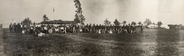 A panoramic group portrait of Ho-Chunk men, women, and children, possibly gathered for a powwow. They are posed standing and sitting in a field in front of some trees, the Dance Lodge, a flag, and various other lodges. The men are wearing pants and shirts while the women wear dresses and embroidered shawls. Near the left side some of the men are holding a large drum.