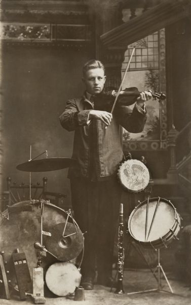 A full-length studio portrait of Hofton C. Hagen posed standing in front of a painted backdrop. He is holding a violin and bow as if he is playing it, and is surrounded by various other musical instruments, including  a drum set, a triangle, and a clarinet.