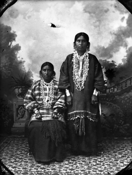 A studio portrait of two Ho-Chunk women in front of a painted backdrop. The woman on the left is posed sitting and is wearing a ribbon work  shirt with silver trader bobs, and jewelry including earrings, necklaces, file bracelets, and rings. The woman on the right is posed standing and is wearing a ribbon work beaded shirt, and jewelry including earrings, necklaces, file bracelets, and rings.