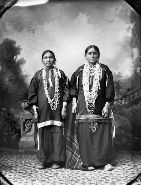 A studio portrait of two Ho-Chunk women wearing similar outfits posed standing in front of a painted backdrop. Both women wear several earrings, necklaces, file bracelets, and moccasins. The woman on the left also wears rings, and a plaid shawl is draped over something in back of them.