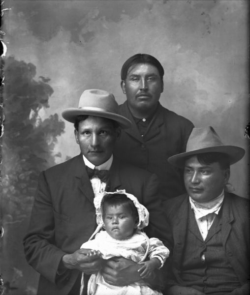 A studio portrait of three Ho-Chunk men and a Ho-Chunk infant in front of a painted backdrop. Two men are posed sitting, and the one on the left is holding the infant, who is wearing a bonnet. Both of the seated men wear hats, and the other man is posed standing behind them in the center. One of the men is identified as the first husband of Mrs. White Gull (or Whitegull).