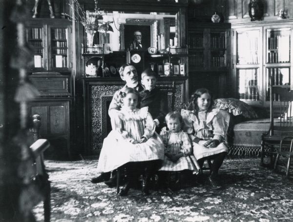 Fred M. Brown holding Eleanore and Timothy III, with Alice and Mary Brown in the foreground in the living room at 121 East Gilman Street.  A portrait of Timothy Brown II is reflected in the mirror over the fireplace.