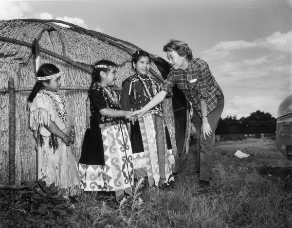 Three young Native American girls meet with a  white woman in front of bark building.