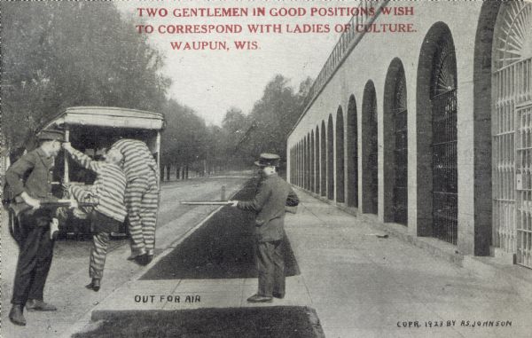 Two police officers holding rifles escort two prisoners into a paddywagon. Lining the road is a long, perspective view of an outdoor prison. Red text imprinted in the upper portion of the image reads, "Two gentlemen in good positions wish to correspond with ladies of culture. Waupun, Wis."