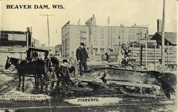 Two men stand staring at a giant pickerel fish laying on a long, wooden horse-drawn sled. The scene takes place in an industrial district, including a lumber yard, a Peerless tobacco building, and some tall electric lines. Text in the upper left corner bears the inscription, "Beaver Dam, Wis."