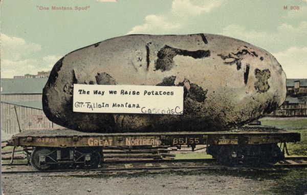 A giant potato rests on a single flatbed railroad car, which is part of the Great Northern line. A white banner on the potato says, "The Way We Raise Potatoes in Montana." Red text in the upper left corner says, "One Montana Spud." Barns or farmhouses stand behind the car.
