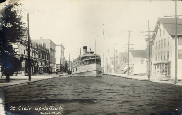 A riverboat travels down a narrow canal. Storefronts, sidewalks and telephone poles line either side of the canal. Caption reads: "St. Clair Up-to-Date."

