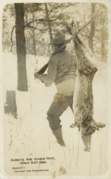 Rear view of a hunter hauling away a giant rabbit through a snowy forest landscape. He is wearing a leather jacket with fringe and boots. Text in the lower left corner bears the words, "Rabbits are scarce here, only got one."