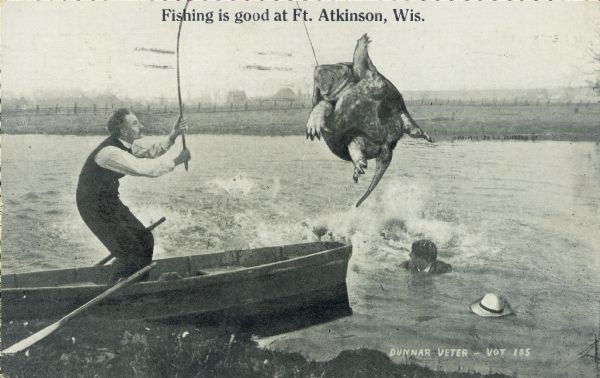 Photomontage of a fisherman in a rowboat catching a giant turtle. The male companion of the fisherman has fallen in and lost his hat amidst the excitement. Farms and a fence are visible across the lake. Black text at the top says "Fishing is good at Ft. Atkinson, Wis."