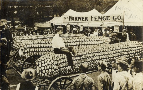 Photomontage of a man, wearing a bow tie and straw boater hat, sitting on top of a giant ear of corn. A crowd of onlookers gather around the man, suggesting that he is attempting to sell this corn. There are three ears in total, and each rests on a flat wooden cart. There is a white tent in the background with a banner in front of it that reads, "The Warner Fence Co." A man holding an umbrella in the background suggests that it may rain soon.