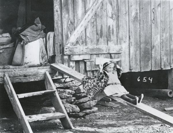 Photographic mock-up for an Alfred Stanley Johnson photomontage postcard entitled "Shooting the Shoots."  A small girl is sliding down a wooden plank, which is resting against the door of a barn.  A short ladder leads up to the door, and inside is a stack of clothes and bags.