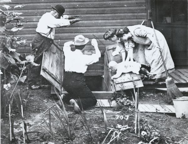 Photographic mock-up for an Alfred Stanley Johnson photomontage postcard entitled "Will It Go In." Two men stand over an open cellar door, with their hands positioned as though they were pushing a large object into it. Two women with brooms are holding a baby by the side of the door.