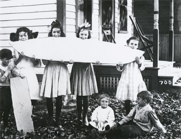 Photographic mock-up for an Alfred Stanley Johnson photomontage postcard entitled "Melon Party."  Four girls wearing bows and dresses are holding an object that resembles a giant slice of watermelon.  A boy stands to the left of them with his hand on a piece of cardboard, while two young children sit on the leaf-covered ground.