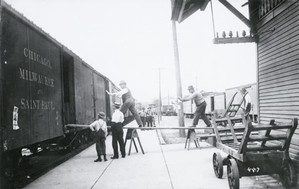 Photographic mock-up for an Alfred Stanley Johnson photomontage postcard. Two men walk up a ramp leading into a railroad car.  Both men are pushing large invisible objects. Johnson would have inserted images of giant vegetables in these vacant spots. The side of the train car says, "Chicago, Milwaukee and Saint Paul."