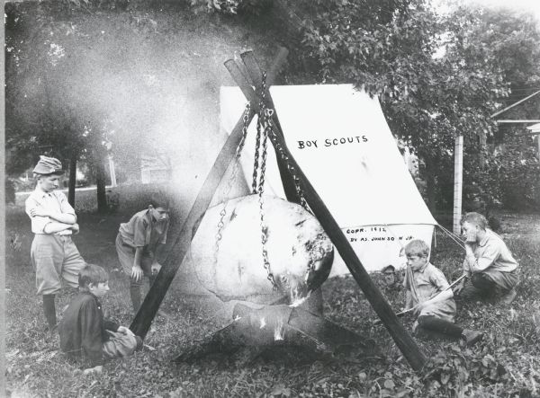 Photomontage of a group of five boy scouts around a cooking fire. The scouts watch as a giant potato, suspended over the fire, slowly roasts. They are wearing uniforms with knee high socks.  They have erected a tent directly behind the fire. The words, "Boy Scouts," have been imprinted on the image as though they were written on the side of the tent.