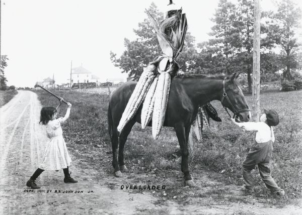 Photomontage of a horse carrying a bundle of giant ears of corn on its back.  A boy is struggling to pull the horse towards the side of the road.  A young girl is about to hit the horse in the tail-end with a stick.  Fences, telephone poles, and farmhouses are visible in the background.  The word, "Overloaded," is imprinted at the bottom of the image field.