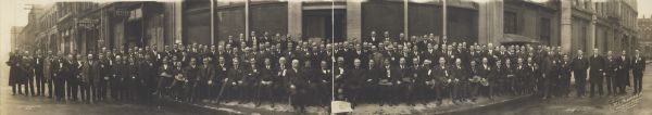 Panoramic view of the Fifth Annual Convention of Western Retail Lumbermen's Association, including the Association members and Lumbermen's Mutual Society. Congress Saloon and Hotel Revere visible in background on street. Boy in front center holding a copy of the "Tacoma Daily News".