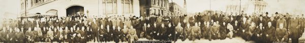 Panoramic group portrait of the 13th annual meeting of the Wisconsin Buttermakers' Association, Feb. 3-5, 1914. Taken outside of the Wisconsin State Capitol. A woman in the center is holding a large hand muff.