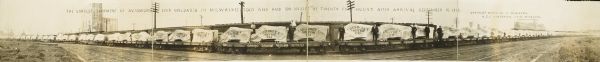 Panoramic view of train loaded with Chevrolet automobiles. Caption at top reads, "The largest shipment of automobiles ever unloaded in Milwaukee, sold and paid for inside of twenty four hours after arrival, September 15, 1916. Chevrolet Motor Company of Milwaukee, H.J.C. Henderson, Manager." Automobiles are covered with sheets branded with "Chevrolet Motor Co." and sit atop Grand Trunk flatbed train cars.
