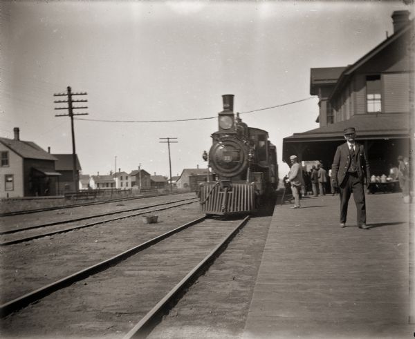View down platform towards locomotive and train at the railroad station. There are people standing on the platform near the depot building.