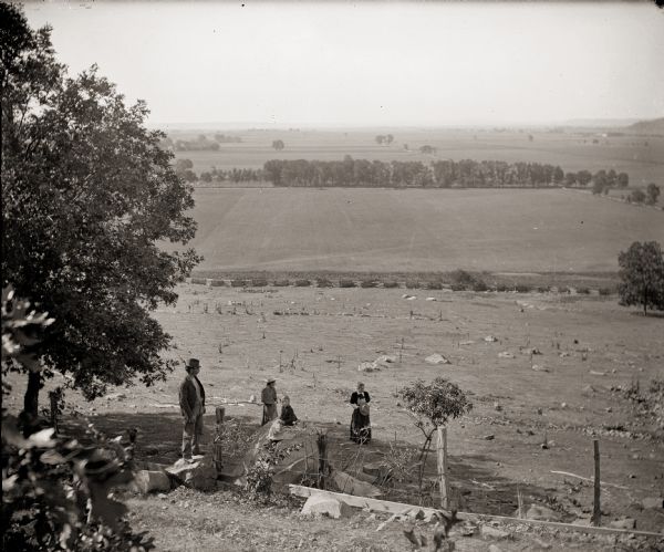 A view of the Sauk Prairie looking south. From the left are the photographer's brother, Frank Bass; Mrs. Edward (Ada Burlingame) Bass and her daughter, Everetta, sitting on boulder; and the photographer's sister, Rhoda Bass. A split rail fence separates the rocky hillside from a field. Headstones in Pioneer Cemetery are just visible beyond the field, on the left. Ferry Bluff is in the background, right. The Badger Ordnance Work later occupied this part of the Sauk Prairie.