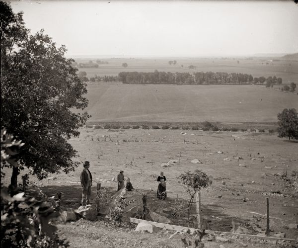 A view of the Sauk Prairie looking south. From the left are the photographer's brother, Frank Bass; Mrs. Edward (Ada Burlingame) Bass and her daughter, Everetta, sitting on boulder; and the photographer's sister, Rhoda Bass.  A split rail fence separates the rocky hillside from a field. Headstones in Pioneer Cemetery are just visible beyond the field, on the left.  Ferry Bluff is in the background, right.  The Badger Ordnance Work later occupied this part of the Sauk Prairie.  