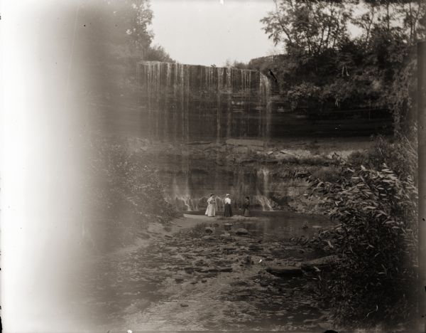 Group of three women, including Ada Bass, at the base of Minnehaha Falls. Minnehaha Creek in foreground.