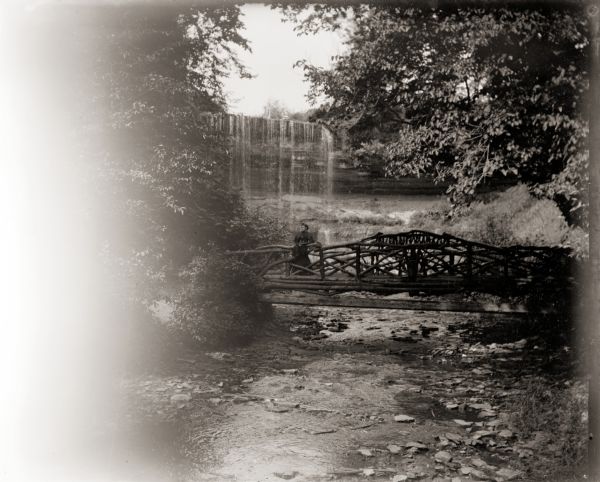 Unidentified woman standing on the footbridge at the base of Minnehaha Falls. Minnehaha Creek in foreground.