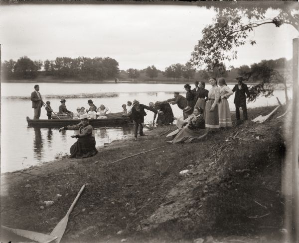 Group of men and women on the shore of an unidentified lake. Nearby, a second group of children, men, and women are pushing off from the shoreline in row boats.