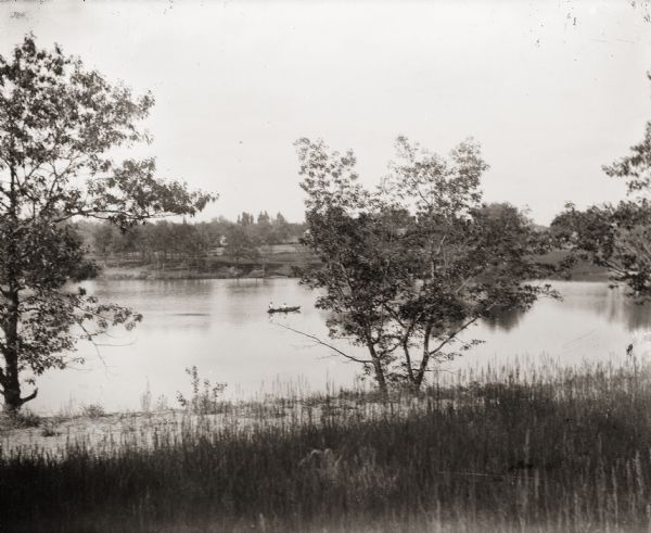 View from shoreline of couple in rowboat on unidentified pond.