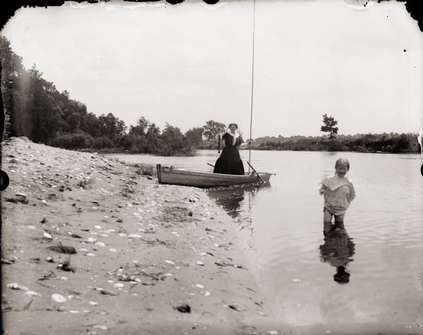 Young girl in foreground, wading in Fox River. Woman standing in a rowboat, pulled to shore, holding a fishing pole and fish.
