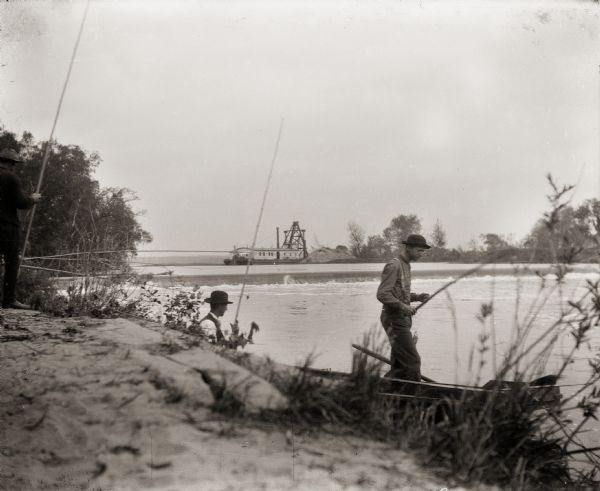 Group of three men fishing on the Fox River, with view of dam. There is a barge in the background.