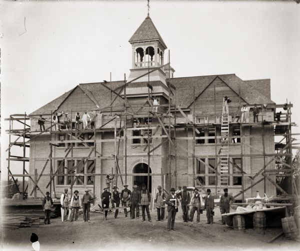 Group of men posing in front of and on the scaffolding surrounding the Montello School House. The building was completed in 1896.