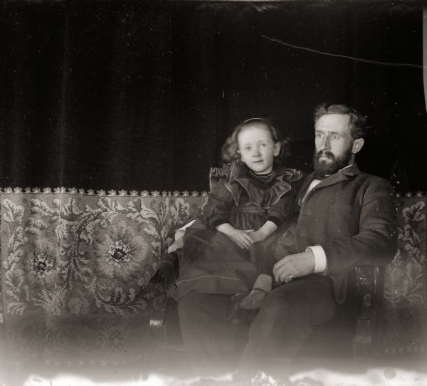 Dr. Edward A. Bass seated, with his daughter Everetta on his knee in front of a curtain.