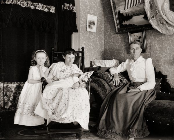 Portrait of Ada Bass seated while holding son Cary, with daughter Everetta standing next to her. Another woman, possibly Ada's mother, Arlina Burlingame, is seated on the adjacent couch.