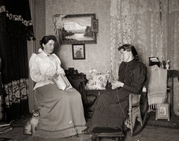 Ada Bass sitting in a rocking chair while crocheting. Sitting across from her is a woman identified as Llora, who is reading. There is a replica of a cat propped at Llora's feet.