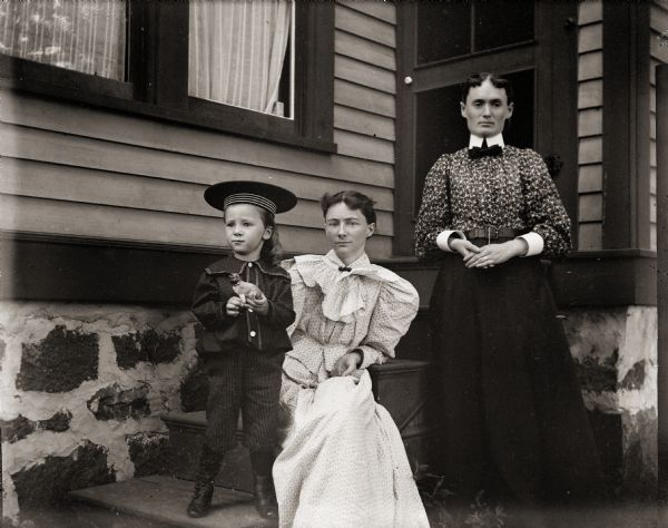 Ada Bass, seated, with son Cary holding a toy dog. Standing next to them is Lizzie Loote. They are on the steps of the Bass family home at 131 East Montello Street.