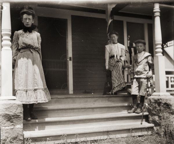 Ada Bass, seated on the porch, with the family dog seated next to her. Her children, Everetta and Cary (holding a cane in his left hand), are standing on the steps of the porch. The photograph was taken at the Bass family home, 207 Clay Street.