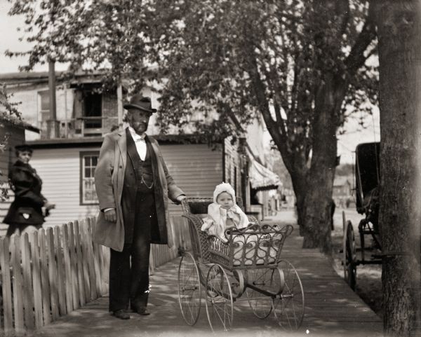 Dr. Edward A. Bass pushing his son Cary in a baby carriage along a wooden sidewalk on Nebraska Street (now West Montello Street).