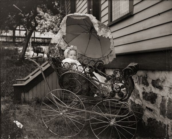 Infant, identified as Loura, seated in a baby carriage outside of the Bass family home, 131 East Montello Street.