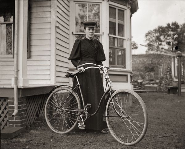Woman identified as Miss Demoth posing with her bicycle near a house. Her bicycle is equipped with a guard around the chain.