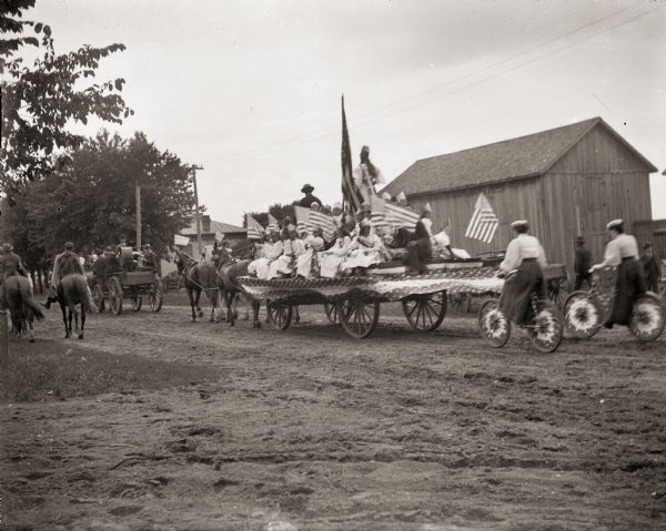 Group of children seated on horse-drawn parade float covered in American flags. They are preceeded by a cart with musicians, and are followed by two women on bicycles.