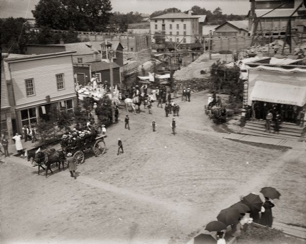 Elevated view of parade turning from Nebraska Street onto Main Street. Women with umbrellas line the sidewalk in the foreground of the image. The Montello Woolen Mill is visible in the center background, with the granite quarry building to the right. The mill pond (Montello Lake), and the Montello River are also seen behind the Woolen Mill.  The awning on the right says "Tonsorial Parlors."